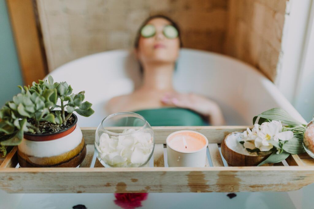 A person relaxes in a bathtub, eyes covered with cucumber slices. A wooden tray holds a candle, succulents, flower petals, and bath salts, conveying tranquility.